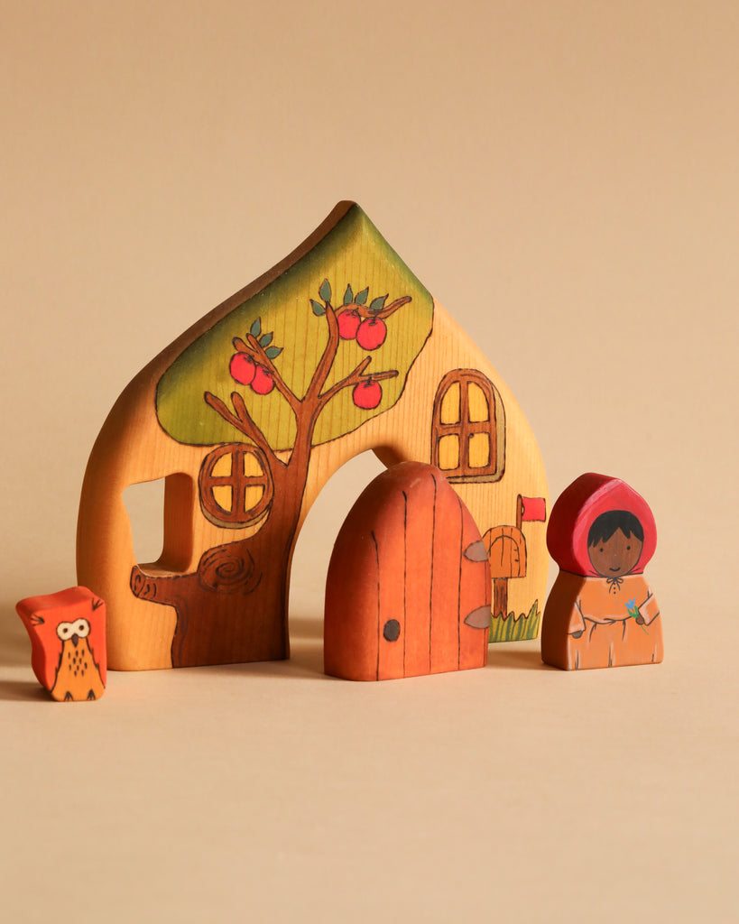 The Handmade Wooden Cottage is a vibrant, handcrafted wooden set that includes a house adorned with a tree and red apples. In front of the house, there is an adorable owl figure and a human figure in a red hooded cloak. Set against a simple beige background, the cottage also features a removable door for extra fun.