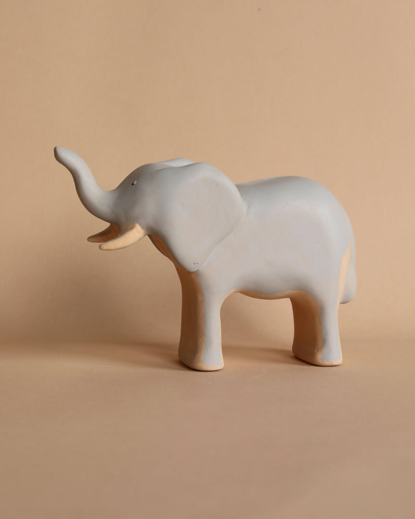 A small, simple Handmade Wooden Elephant stands on a neutral beige background. The elephant, with its trunk raised, has a minimalist design with smooth surfaces and light detailing, resembling handcrafted toys, giving it a serene appearance.