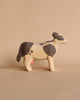 A Handmade Wooden Cow with white and brown spots stands on a neutral beige surface with a beige background. Handmade in Serbia from linden wood and featuring child-safe paint, the cow has a simplistic and rustic design with minimal details, including a pink udder.