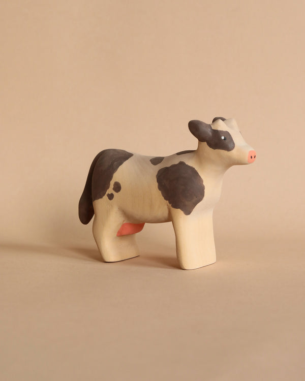 A Handmade Wooden Cow, handcrafted from linden wood, stands on a tan surface. Painted with black and white patches using child-safe paint, the figurine features a charming design with a pink udder and a minimalistic facial expression. Handmade in Serbia, the background is a solid beige color.