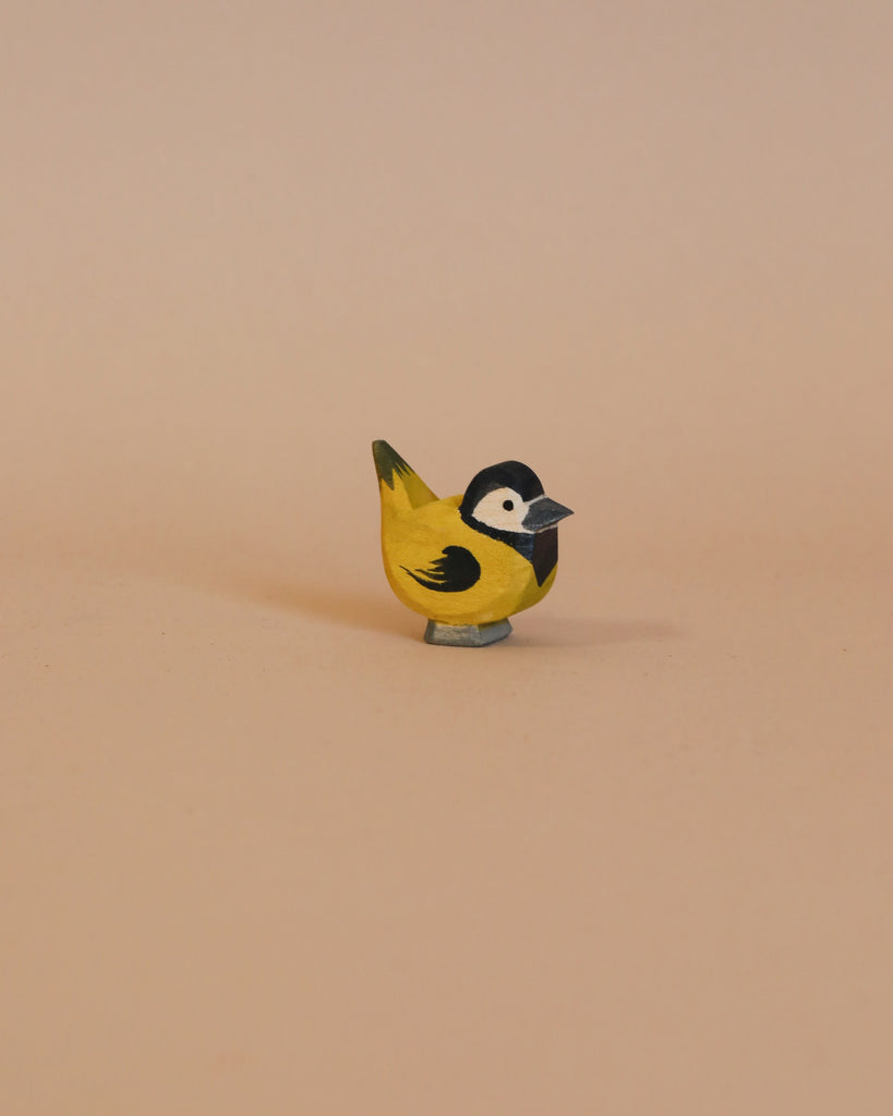 The Ostheimer Goldfinch is a small, handcrafted wooden bird figurine made from sustainably sourced materials. It is painted in yellow, black, and grey tones and features a black head with white cheeks and a yellow body, closely resembling a great tit. The bird is shown against a neutral light beige background.