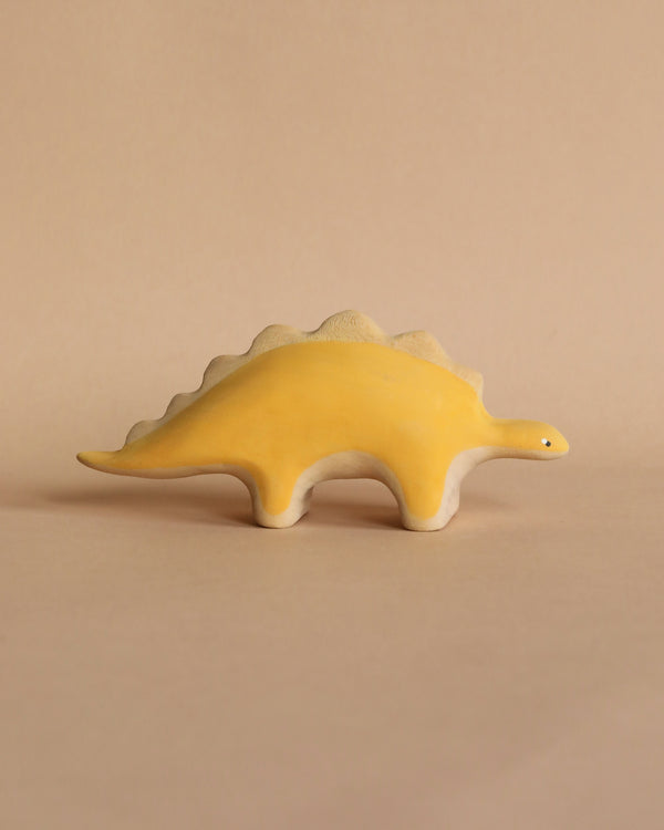 A Handmade Wooden Stegosaurus Dinosaur, specifically a handcrafted wooden stegosaurus, with a simple and smooth design. It is painted yellow with non-toxic paint and features beige accents on its underside. The toy stands on a beige surface with a matching beige background.