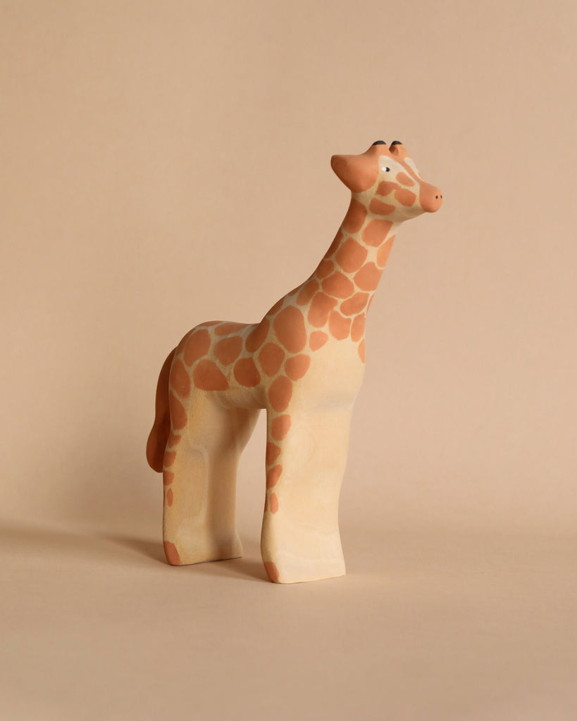 A small, simplistic Handmade Wooden Giraffe with orange and white spots stands on a beige surface against a beige background. Handmade in Serbia, the giraffe has an elongated neck and slightly wide legs, resembling a children's toy or ornament.