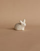 A small, white, minimalist handmade wooden rabbit, made in Serbia, is positioned against a plain, light beige background. The figurine has simple black dot eyes and a tiny pink nose and is shaped in a rounded, smooth form with little detail.