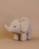 The Elephant Teddy Stuffed Animal, in light beige with a small black eye, stands against a plain, light brown background. Its fabric texture mimics soft, curly fleece, while the plush toy's polyester filling makes it extra cozy.