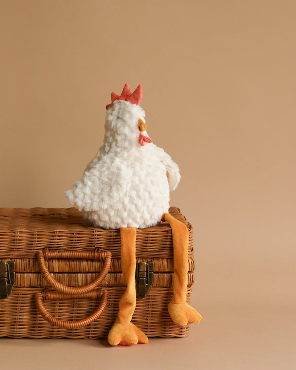 A fluffy white Charlie the Chicken Stuffed Animal, featuring a red comb and long yellow legs, sits on a woven wicker basket against a beige background. Charlie's legs dangle over the edge of the basket.