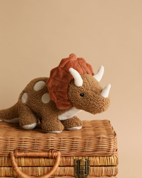 The Triceratops Dinosaur Stuffed Animal, featuring a brown body with white spots and a rust-colored frill, stands on a closed wicker basket secured with a clasp. Measuring 40cm x 18cm, it is suitable for ages 0+ and can be cleaned using a cold gentle machine wash. The background is a plain beige wall.