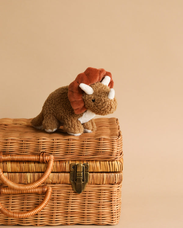 Meet the Baby Triceratops Dinosaur Stuffed Animal, a charming triceratops toy with a brown body and a reddish-brown frill, sitting adorably on a closed wicker basket against a beige background. Perfect for tiny dino enthusiasts, this plush toy is suitable for infants.