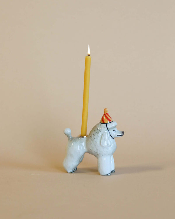 A whimsical Poodle Cake Topper with a yellow candle lit on its back, set against a plain beige background. The poodle wears a small, festive red hat with gold trim.