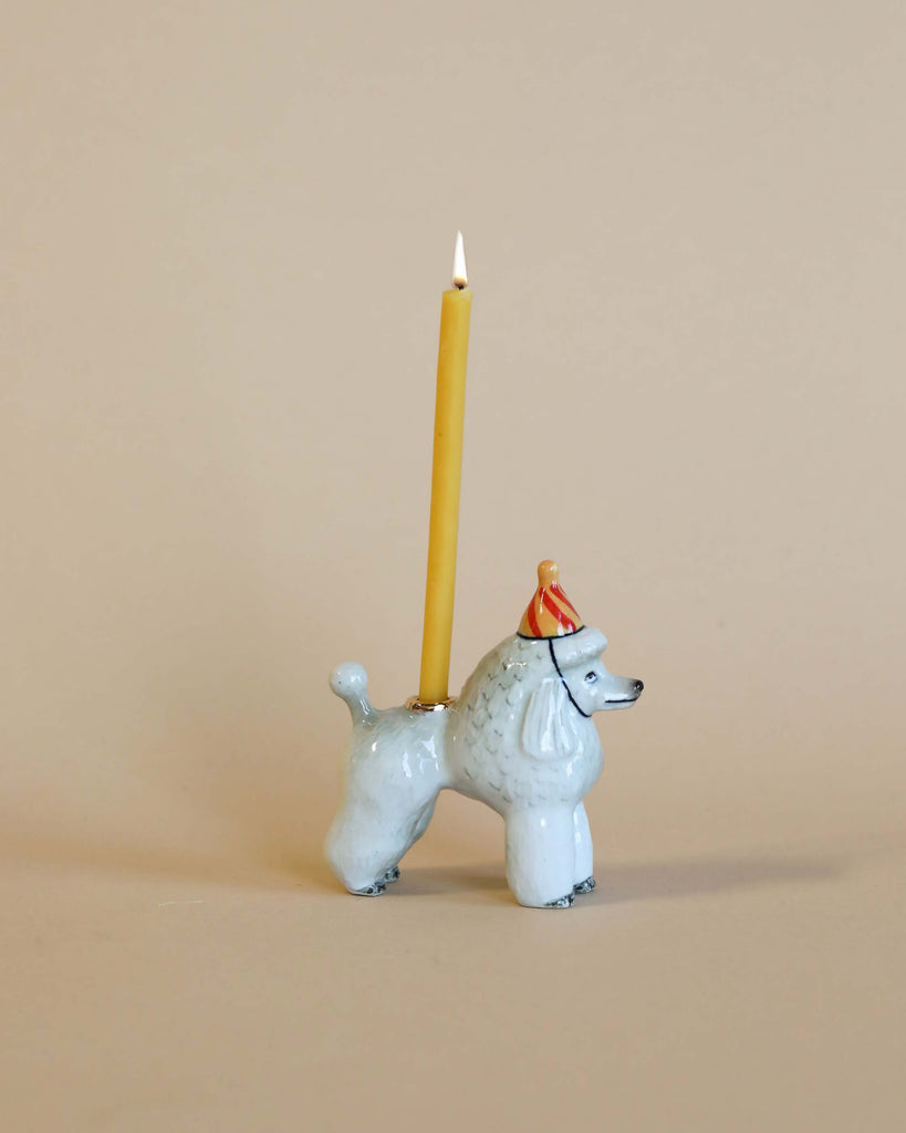 A whimsical Poodle Cake Topper with a yellow candle lit on its back, set against a plain beige background. The poodle wears a small, festive red hat with gold trim.