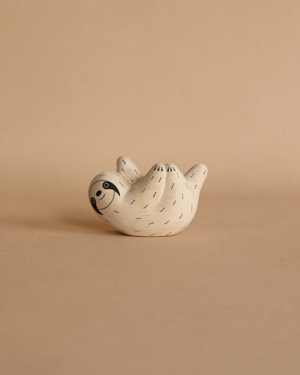 A small, white ceramic figurine of a sloth lies on its back with a smiling face, set against a beige background. The sloth has simple black lines indicating fur and expressive eyes, giving it a whimsical appearance, reminiscent of the Handmade Tiny Wooden Forest Animals - Sloth.