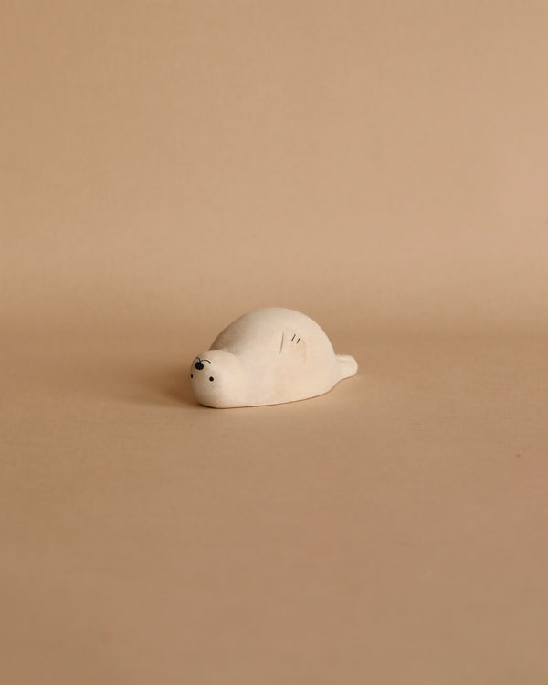 A small, Handmade Tiny Wooden Seal lies on its belly against a plain beige background. Hand-carved from eco-responsible forests, the seal's minimalistic design features tiny black dots for eyes and nose, with subtle line details on its back, all contributing to its cute and simplistic appearance.