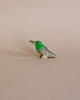 A small Handmade Holzwald Kolibri Bird figurine with a green head and grey body, set against a plain light beige background, epitomizes sustainable wooden toys.