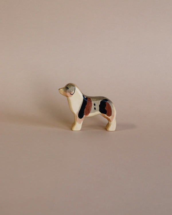 A Handmade Holzwald Australian Shepherd, painted with patches of black, white, and brown, standing against a plain light beige background. The dog appears in a stationary, profile pose.