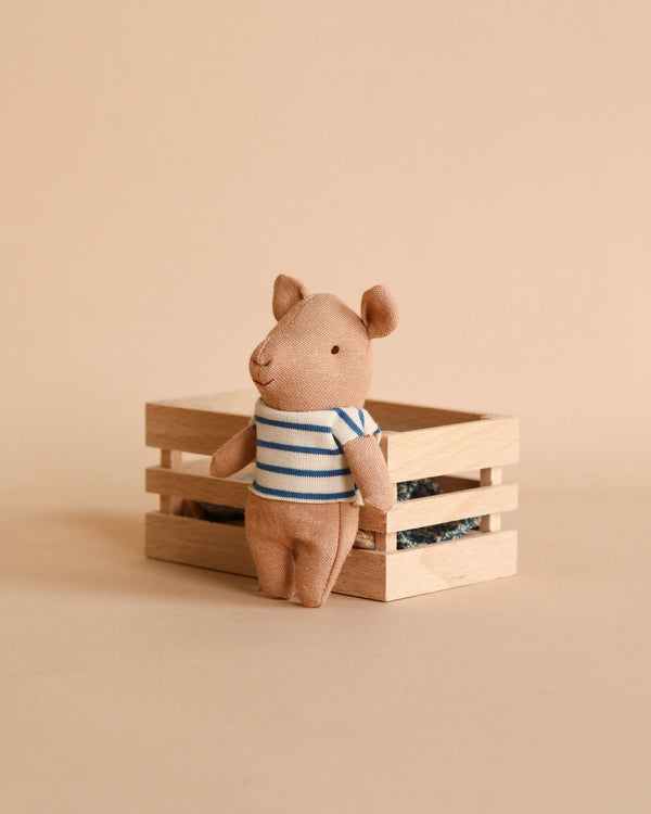 A small, plush Maileg Pig In Box, Baby Boy - Blue toy wearing a blue-and-white striped shirt stands posed against a light beige backdrop. The pig is leaning slightly on a simple wooden box placed behind it, creating a cute, whimsical scene.