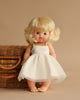 A Minikane Baby Doll (13") named Angela, with blonde hair and blue eyes, stands in front of a wicker basket. Though she's barefoot and dressed in cream-colored clothing that is sold separately, this anatomically correct doll exudes simple charm against the plain beige background.