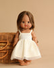 A Minikane Baby Doll (13") - Leopoldine, with long brown hair, stands barefoot in front of a wicker basket. She's dressed in a sleeveless white dress, and the neutral beige background enhances the simplicity and elegance of the scene. A subtle natural vanilla scent adds to its charming allure.