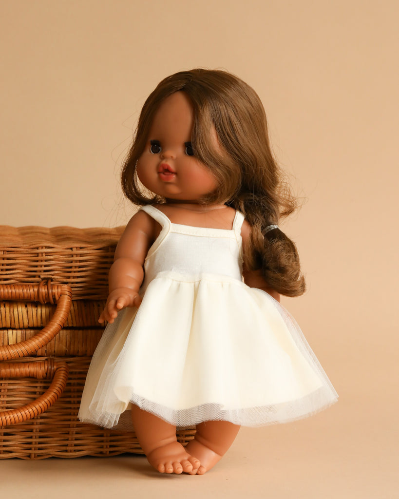 A Minikane Baby Doll (13") - Leopoldine with long brown hair and dark skin is standing next to a wicker basket. The doll, wearing a white sleeveless dress and exuding a natural vanilla scent, has a loose braid over one shoulder. The background is a plain beige color.