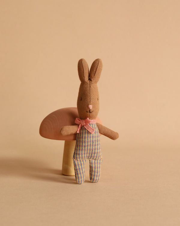 A small, Maileg My Rabbit - Rose/Blue stands upright against a beige background, wearing a blue and white checkered jumpsuit with a pink bow at the neck. Behind the rabbit, there is a wooden mushroom decoration. This must-have accessory for any nursery table evokes a soft, neutral tone.