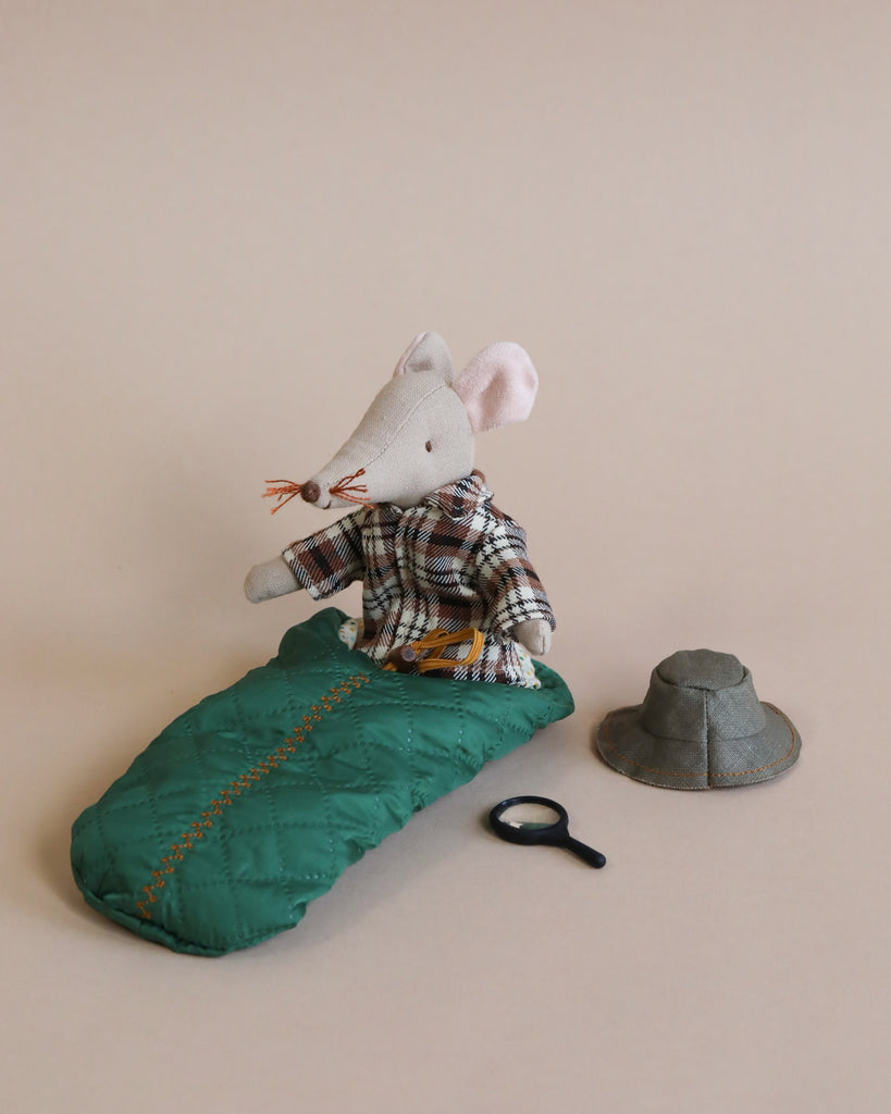 A Maileg Wildlife Guide Mouse dressed in a plaid shirt and overalls is sitting atop a green sleeping bag, next to a gray felt hat and a black magnifying glass. The background is pale beige.