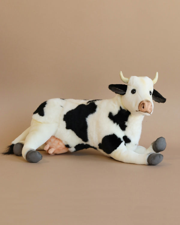 A Cow Stuffed Animal, featuring white and black spots, is lying down against a plain beige background. This artisan hand-sewn plush toy from our collection has small yellow horns, a pink udder, and grey hooves. Its ears and tail are black, matching some of the spots on its body.