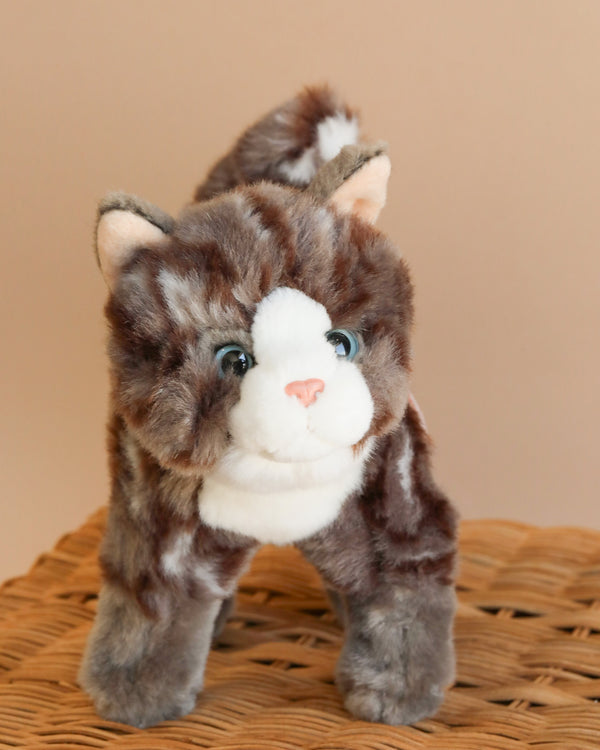 The Teddy Hermann Standing Tabby Cat Stuffed Animal, featuring brown, white, and grey fur with blue eyes, stands on a woven wicker surface against a beige background. This high-quality plush toy boasts a friendly and happy expression.