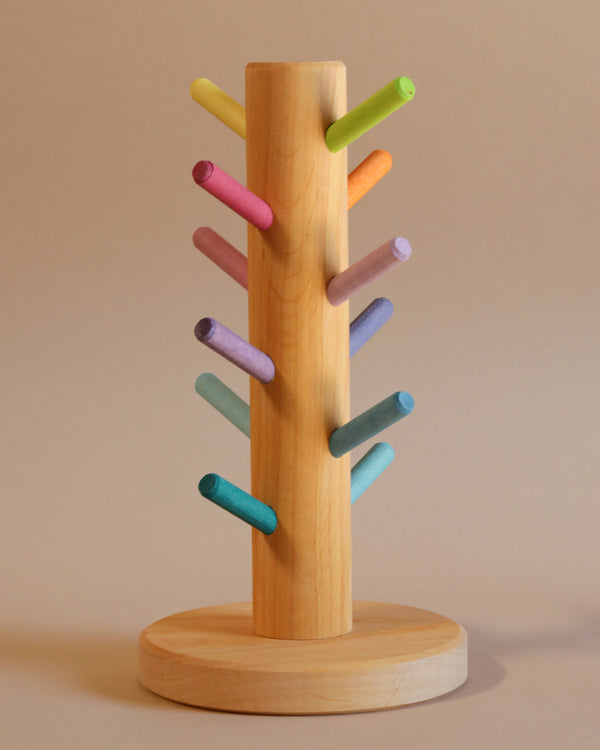 The Grimm's Pastel Sorting Helper for Building Rings is a wooden dowel stand featuring pastel-colored pegs extending from it at various angles. The pegs are painted in lovely shades of yellow, pink, red, purple, blue, and green. The stand boasts a round and smooth base that is complemented by wooden discs. All of this is set against a plain beige background.