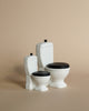 A minimalist image featuring a bathroom assortment of three small, white porcelain objects resembling fixtures: a Maileg Miniature Toilet - Teddy Size, a tall toilet with a vertical tank, and a urinal. Each has black accents and is placed against a beige background.