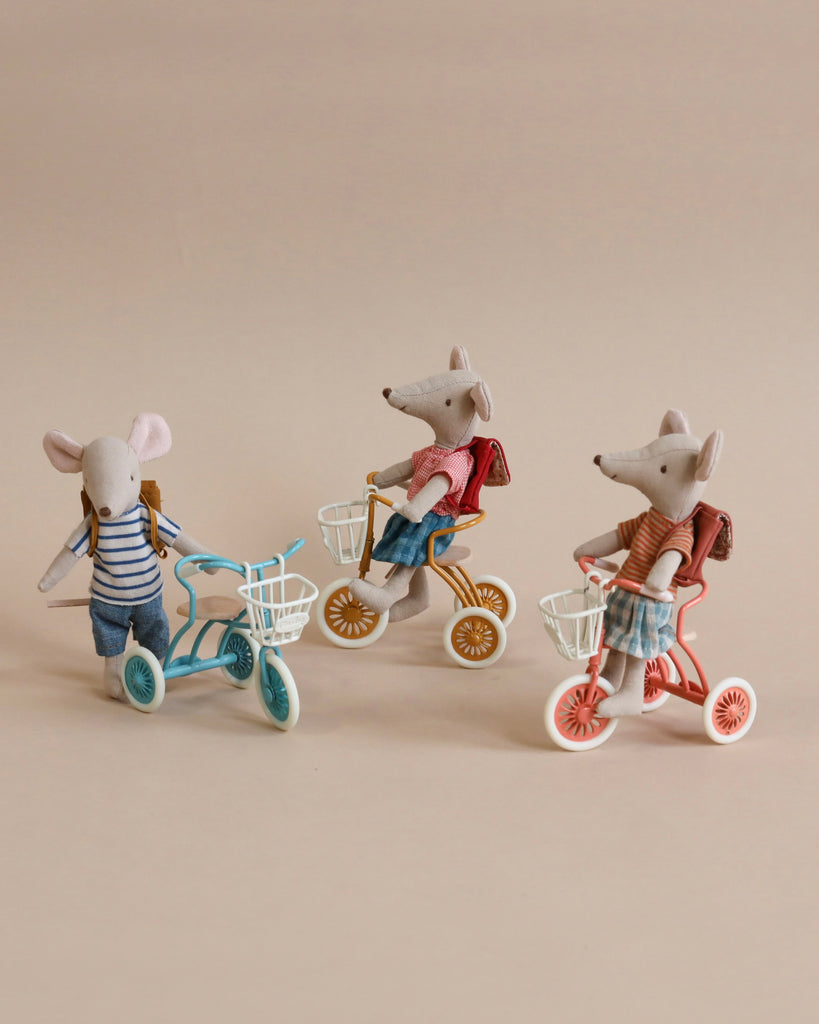 Three Maileg Tricycle With Baskets, each with magnetic hands and wearing backpacks, are riding colorful toy bicycles. One mouse wears a striped shirt and rides a blue bike with wooden seats, while the other two wear different outfits and ride orange and red bikes, all against a plain beige background.