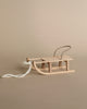 A small, wooden Maileg Christmas Mini Sled with a metal handlebar and a white rope attached to the front is placed on a neutral, beige background. This winter accessory features a simple, rustic design.