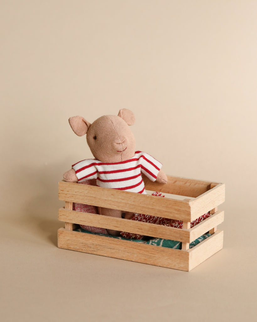 A small, stuffed animal resembling a pig with a light pinkish-brown body is dressed in a red and white striped shirt. It is sitting inside a wooden box that resembles a playset with some folded fabric pieces. The background is a neutral beige color.