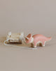 A handcrafted Handmade Wooden Triceratops Dinosaur Push Toy With Built-In Magnets featuring a pink triceratops and a rectangular cart. The triceratops has a string attached to it that connects to the cart. Both the dinosaur and the cart are made of light-colored wood, with the triceratops partly painted pink using non-toxic paint.