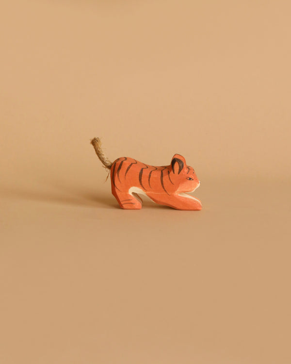 A small handcrafted wooden figurine of an orange tiger with black stripes and a rope tail, named Ostheimer Tiger Lurking, Small, is placed against a beige background. The toy appears to be in a crouching or playful position, perfect for imaginative play.