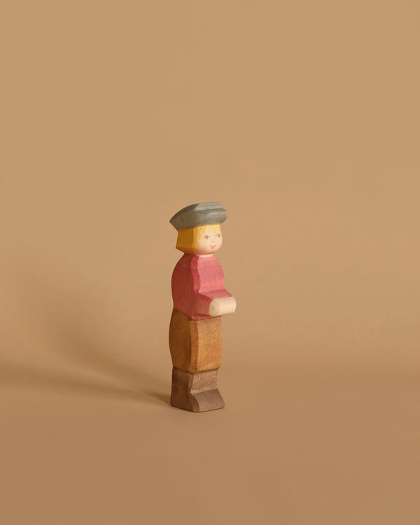 An Ostheimer Son, Gray Hat wooden toy figure with simplistic features stands against a beige background. It has blonde hair, a light gray hat, a red shirt, light brown pants, and brown shoes. Perfect for imaginative play, the handcrafted figure has a neutral expression and faces slightly to the right.