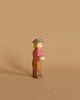 An Ostheimer Son, Gray Hat wooden toy figure with simplistic features stands against a beige background. It has blonde hair, a light gray hat, a red shirt, light brown pants, and brown shoes. Perfect for imaginative play, the handcrafted figure has a neutral expression and faces slightly to the right.