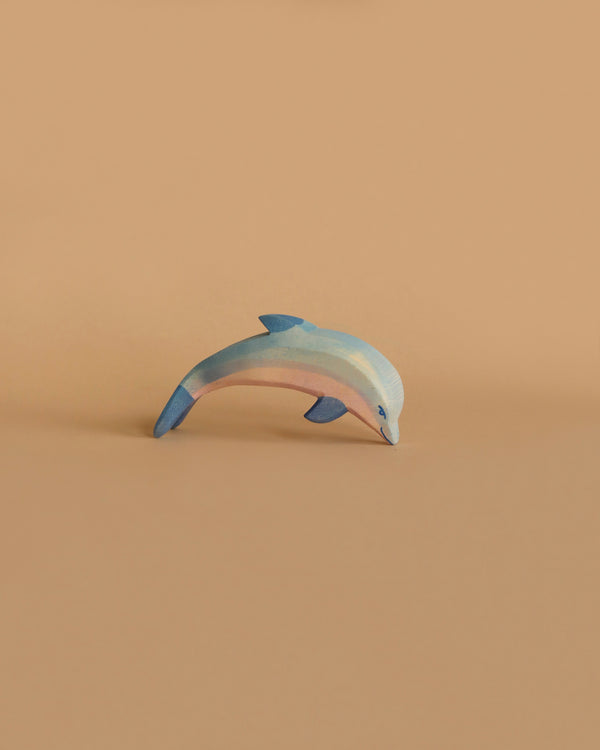 The Ostheimer Dolphin, Head Low figurine is small and smooth, handcrafted from wood with shades of blue and white. Positioned as if in a playful, leaping stance, it stands out against an orange-brown background.