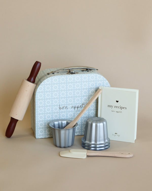 A small baking kit for tiny bakers is displayed, featuring a decorative box, wooden rolling pin, spatula, metallic molds, wooden spoon, and a recipe book titled "my recipes." The Madeline Baking Set - Petite Cousine items are arranged neatly on a neutral background.