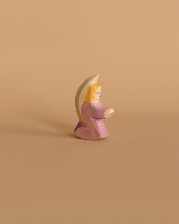 The Ostheimer Little Angel - Purple is a handcrafted wooden figurine depicting an angel with light brown hair, dressed in a lilac robe, holding a crescent moon behind its back. Made from sustainably sourced materials, this minimalist piece evokes the enduring charm of Ostheimer wooden toys and is beautifully set against a white background.