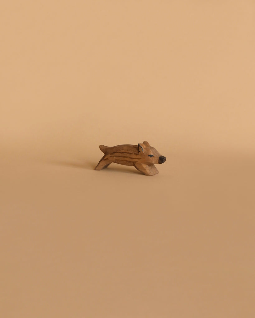 A small, light brown wooden figurine of a wild boar in a playful stance, handcrafted by Ostheimer, placed on a beige background.