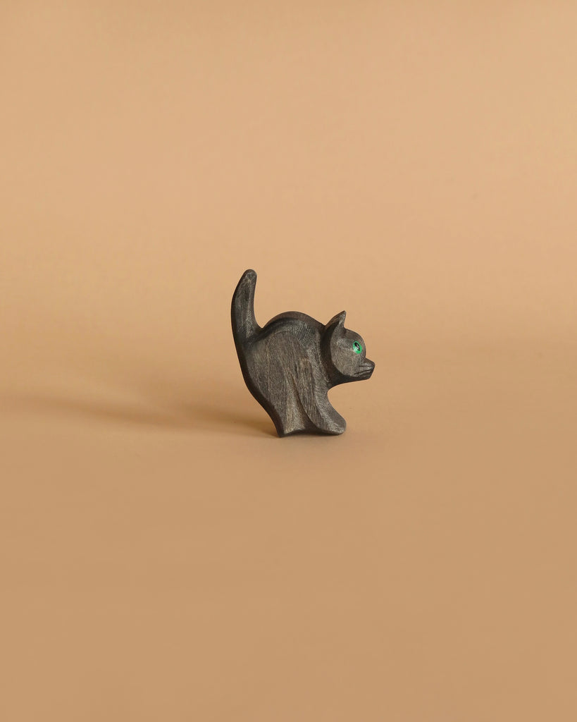 The Ostheimer Cat For Witch, a small, dark wooden figurine with an upturned tail and captivating emerald green eyes, stands against a plain, light brown background. This charming piece evokes the essence of handcrafted wooden toys that are perfect for imaginative play.