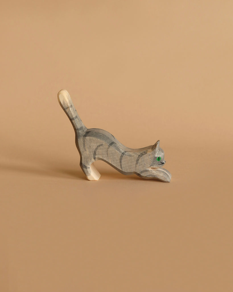 The Ostheimer Gray Cat, Jumping is a small carved wooden figurine, painted gray with darker stripes and depicted in a playful, stretching pose. The cat features green eyes that evoke imaginative play and is positioned on a solid beige background, showcasing the handcrafted quality characteristic of Ostheimer toys.