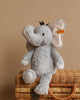 A Steiff Ella Elephant Plush Animal Toy, standing 12 inches tall, features soft gray fur, large floppy ears lined in pale pink fabric, black eyes, and a black tuft of hair on top of its head. It sits upright with a small Steiff tag attached to one ear.