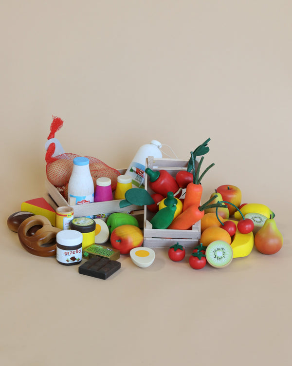 A variety of toy food items are arranged on a light brown surface. The collection includes plastic fruits, vegetables, dairy products, eggs, chocolate, and bread—all colorful and detailed. Perfect for a pretend play store, the pieces are organized in small wooden crates and scattered around them. This is the Erzi Big Box of Play Food Shop Assortment.