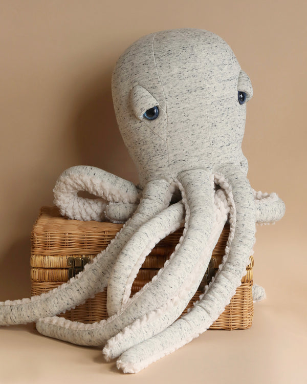 The BigStuffed Octopus - Big Original, featuring a soft grey fabric body and long tentacles, rests on a wicker basket against a beige background. The octopus has large, round eyes and a calm expression, perfectly capturing the charm of the underwater world.