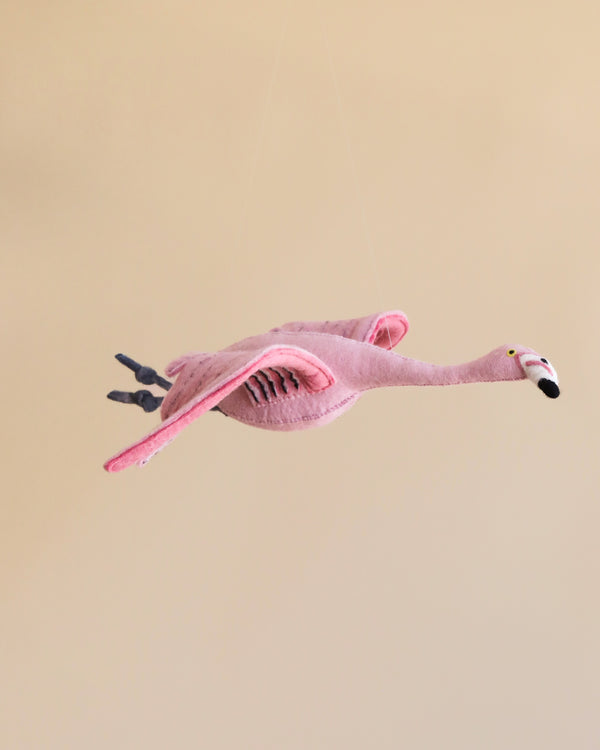 The Hanging Felt Flamingo is suspended in mid-air against a neutral beige background. Made from organic wool and detailed with eco-friendly dyes, the pink flamingo features outstretched wings, black and white accents on its beak, as well as grey legs and feet. Strings give the illusion that it is flying.