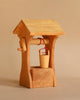 The Ostheimer Well with Roof is a small, hand-carved wooden well featuring a pitched roof and a simple crank handle. A tiny wooden bucket, suspended by a red string, hangs from the central beam. Handcrafted using sustainable materials, this charming piece is perfect for imaginative play and stands on a plain, neutral background.