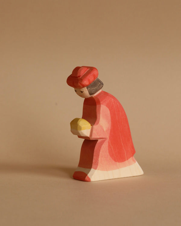 The Ostheimer Red King is a small, wooden figurine of a person wearing a red hat and red coat, and holding a yellow object. Handcrafted from sustainably sourced materials, this simple, minimalist toy is set against a solid beige background.