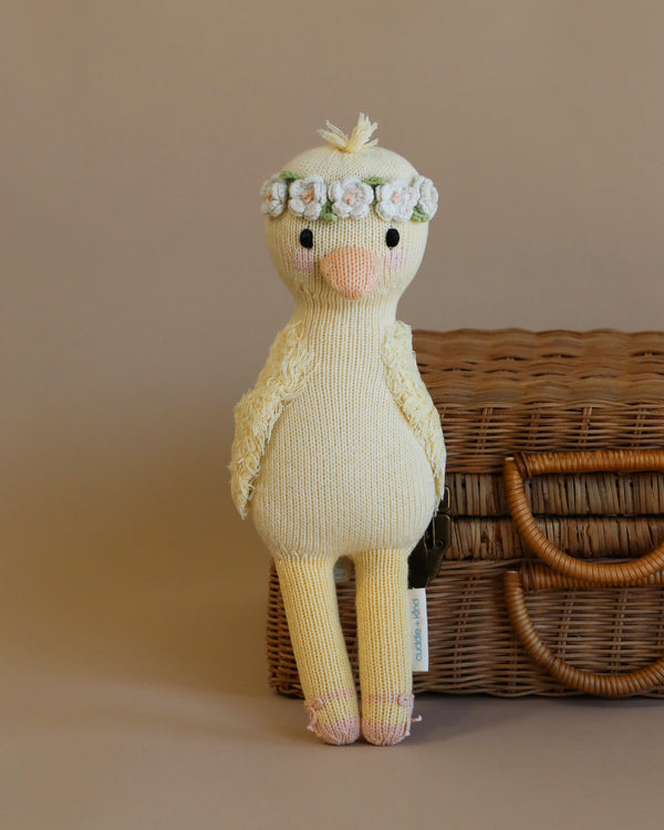 A Cuddle + Kind Duckling with a floral crown, standing in front of a wicker basket against a plain beige background. The duckling, filled with hypoallergenic polyfill, features detailed stitching.