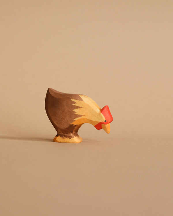 The Ostheimer Hen Picking - Brown, a small wooden figurine handcrafted from sustainably sourced materials and painted in natural brown and beige shades with a bright red comb, is displayed against a plain beige background. The hen appears to be pecking at the ground.