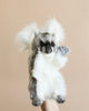 A hand is holding the Koala Puppet against a beige background. The fluffy, gray and white plush toy features large, fluffy ears, a dark nose, and small black eyes, giving it cute and realistic characteristics.